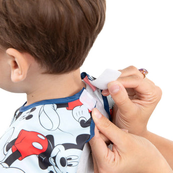 An adult ties a waterproof Bumkins Junior Bib: Mickey Mouse Classic, featuring easy-wipe fabric, around a childs neck.