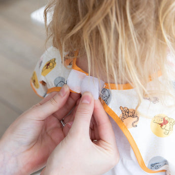 Person fastening the Velcro on a colorful Bumkins Junior Bib: Winnie and Friends, with playful animal designs.