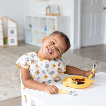 A child in a patterned shirt wears a colorful Bumkins Junior Bib: Winnie and Friends at a table with food on a yellow plate.