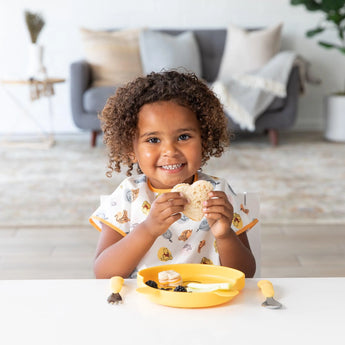 A child beams, holding a sandwich with a yellow plate and utensils, wearing a Bumkins Junior Bib: Winnie and Friends in the cozy living room.