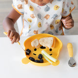 A child eats sandwiches, blackberries, and celery from a bear-shaped yellow plate while wearing a Bumkins Junior Bib: Winnie and Friends.