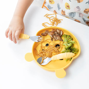 A childs hand with a fork near a bear-shaped plate of food, complemented by a Bumkins Junior Bib: Winnie and Friends on a white table.