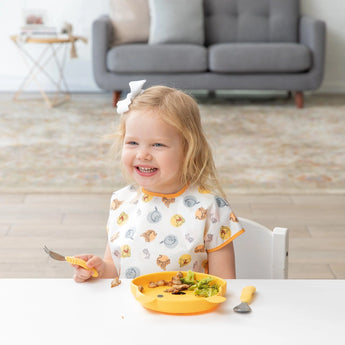 A girl in a patterned shirt smiles while eating with a fork from a yellow plate wearing a Bumkins Junior Bib: Winnie and Friends.