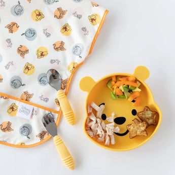 Bumkins Junior Bib: Winnie and Friends baby plate set includes food, matching fork and spoon on a patterned cloth.