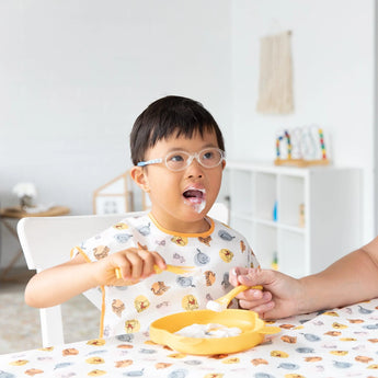 A child with glasses and a print shirt enjoys a meal with an adult nearby, wearing Bumkins Junior Bib: Winnie and Friends.