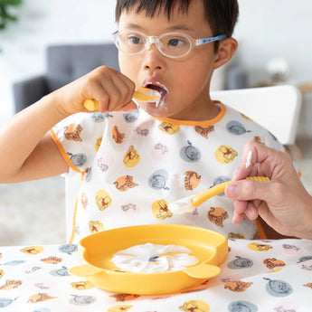 A child in glasses wears a Bumkins Junior Bib: Winnie and Friends while eating yogurt from a yellow bowl with adult assistance.