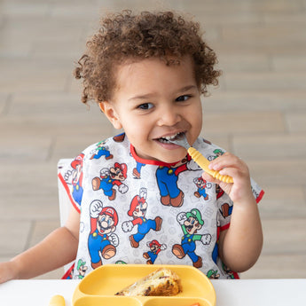 A curly-haired child in a Bumkins Super Mario™ & Luigi bib eats from a yellow plate, smiling brightly with a fork held up.