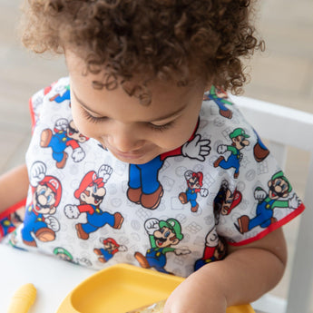 A curly-haired child enjoys a meal with a yellow tray, wearing a colorful Super Mario™ & Luigi Junior Bib by Bumkins.