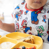 A child in a colorful Bumkins Junior Bib: Super Mario™ & Luigi holds a fork near a yellow divided plate with food.