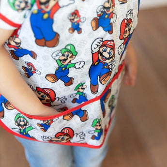 Child in Bumkins Junior Bib: Super Mario™ & Luigi and a white shirt with colorful cartoon print, standing indoors.