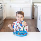 A child happily eats off a blue divided plate, wearing a Bumkins Mario & Luigi bib, with colorful food in a kitchen setting.