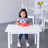 A child in a Bumkins Super Mario™ & Luigi bib smiles at a white table, enjoying food on a pink plate with flowers and white chairs nearby.