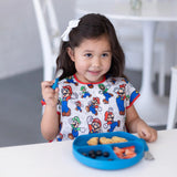 Young girl with dark hair in a bow wearing a Bumkins Super Mario™ & Luigi bib, holding a spoon, sits with her blue plate of food.