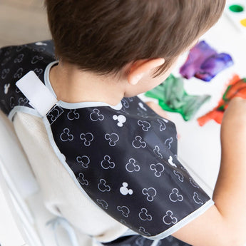 Child painting while wearing a Bumkins short-sleeved smock with Mickey Mouse icons in black and white. Adjustable tie waist.