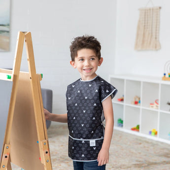 A boy in a Short-Sleeved Mickey Mouse Icon B+W smock by Bumkins stands by an easel, smiling and ready to paint in the playroom.