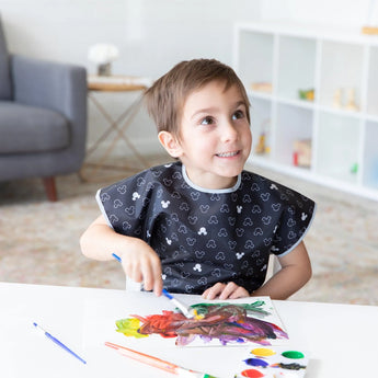 A child painting with watercolors, wearing a Bumkins Mickey Mouse Icon B+W smock in a cozy room.