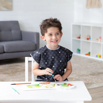 A child paints in a bright room, wearing a Bumkins Mickey Mouse Icon B+W smock, near a gray sofa and shelf.