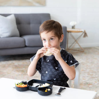 A child wearing a Mickey Mouse B+W short-sleeved smock by Bumkins enjoys a sandwich with food on a sectioned tray, cozy sofa behind.