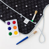 Art supplies on a white background: two paintbrushes, a tray of six vibrant paints, and a Bumkins Short-Sleeved Smock featuring small Mickey Mouse head patterns in B+W. Its crafted from waterproof fabric for easy cleaning after creative adventures.