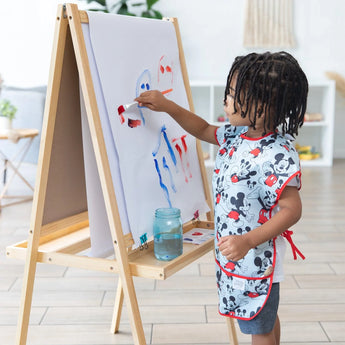 A child in a Bumkins Mickey Mouse Classic smock paints vibrant strokes at an easel on wooden floors in a bright room.