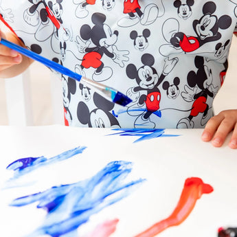 A child painting in blue, wearing a Bumkins Mickey Mouse Classic smock, adds creativity to their colorful day.