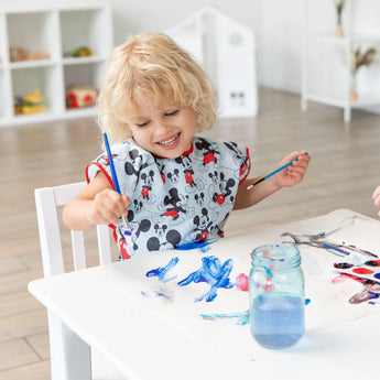 A child paints at a white table in a bright room, wearing a Bumkins Mickey Mouse Classic short-sleeved smock, their blonde hair gleaming.