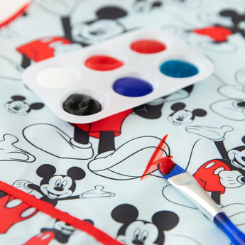 A paintbrush with red paint lies on a Mickey Mouse-patterned surface beside a vibrant palette and a Bumkins Mickey Mouse Classic Smock.