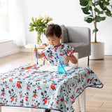 A child paints at a table in a Mickey Mouse Classic smock by Bumkins, amidst blue water, art supplies, and a potted plant.