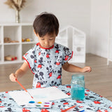 A child paints wearing a Bumkins Mickey Mouse Classic short-sleeved smock, with paints and a jar of blue water on the table.