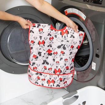 Someone holds a waterproof Bumkins Short-Sleeved Smock: Minnie Mouse Classic near a washing machine.