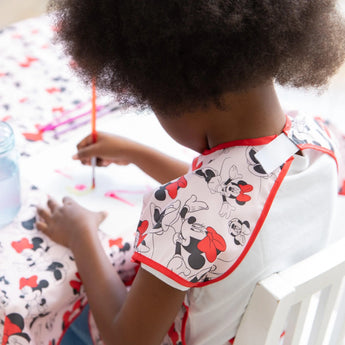 A child paints at a table with Minnie Mouse fabric, wearing Bumkins Short-Sleeved Smock: Minnie Mouse Classic with adjustable tie waist.