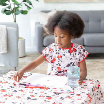 A child draws at a table with Minnie Mouse items, wearing Bumkins Short-Sleeved Smock: Minnie Mouse Classic, surrounded by markers.