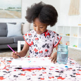 A young curly-haired girl paints at a table wearing a Bumkins Minnie Mouse Classic smock, with a brush and water jar nearby.