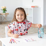 A child smiles while painting in a Bumkins Minnie Mouse Classic smock, with art supplies scattered on the white table.