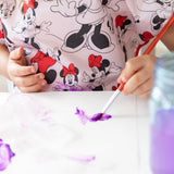 Child painting with purple paint wearing a Bumkins Minnie Mouse Classic smock, featuring an adjustable tie waist, hands and brush in focus.