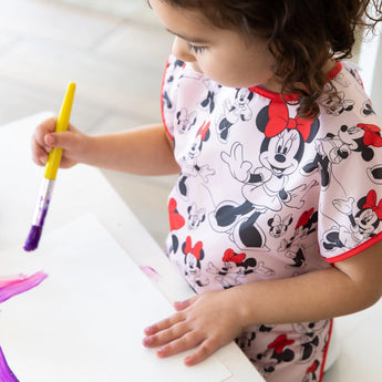 Child painting with a purple brush, in a Bumkins short-sleeved, waterproof Minnie Mouse Classic smock with an adjustable tie waist.