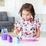 A child wears a Bumkins Minnie Mouse Classic smock, joyfully painting with colorful paints at a table in a bright room.