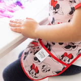 A child in a Bumkins Minnie Mouse Classic Short-Sleeved Smock sits at the table, hands on its waterproof fabric.