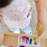 A child paints joyfully with colorful paints, protected by their Bumkins Short-Sleeved Smock: Princess Magic for easy cleaning.