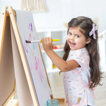 A smiling young girl with long brown hair paints at an easel, wearing a Bumkins Short-Sleeved Smock: Princess Magic and a hair bow.