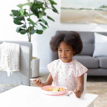 A child with curly hair enjoys a meal from a pink plate, wearing the Bumkins Short-Sleeved Smock: Princess Magic in the bright living room.