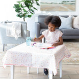 In a cozy living room, a child in their Bumkins Princess Magic Smock paints at a table with supplies and a patterned cloth.