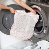 A person places a Bumkins Short-Sleeved Smock: Princess Magic in a front-loading washer, with a nearby laundry basket for easy cleaning.