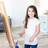 A young girl beams as she paints, her Bumkins Short-Sleeved Smock: Princess Magic ensuring easy cleanup and fun creativity.