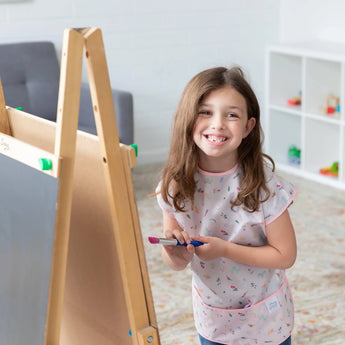 A girl with a paintbrush smiles in a bright room, wearing Bumkins Short-Sleeved Smock: Princess Magic.