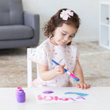 A young girl with a pink bow paints at a small table in a patterned dress and Bumkins Princess Magic Smock; purple paint jar nearby.