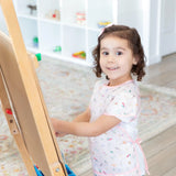 A young girl smiles at an easel wearing a Bumkins Short-Sleeved Smock: Princess Magic, the waterproof fabric shining brightly.