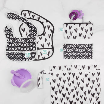 A Bumkins SuperBib, Wet Bag: Hearts, plus a purple cup and teether are on the white surface.