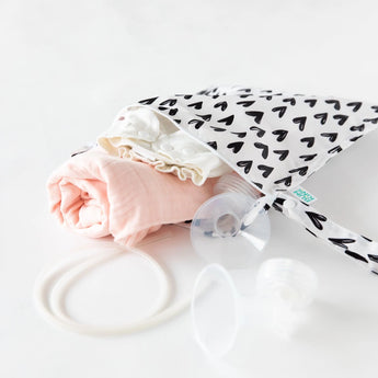 A baby care kit with a pink blanket, white clothes, and Bumkins Wet Bag: Hearts in waterproof fabric.