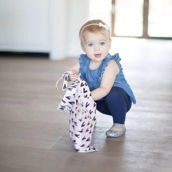 A toddler in a blue outfit crouches, clutching a Bumkins Wet Bag with a heart pattern.